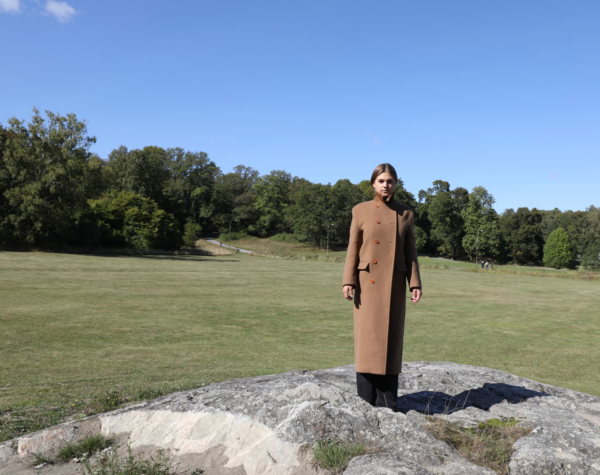 A woman in a camel coat standing on a rock in an open green park