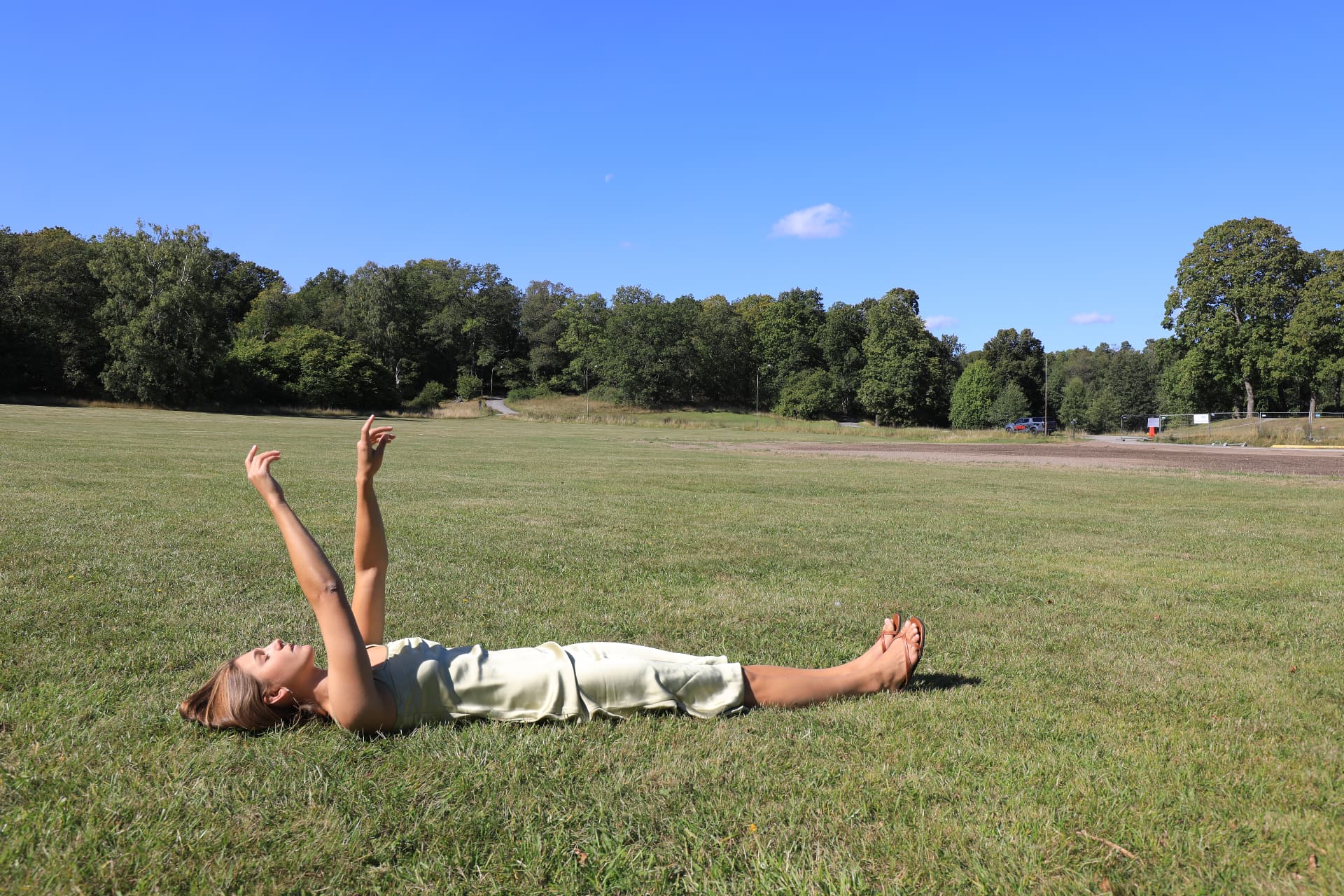 A woman in a pale dress lying in a wide grass field with her arms reaching toward the sky